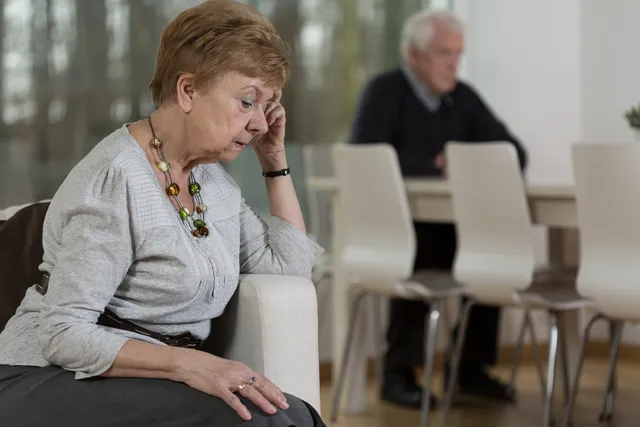 Worried elderly woman with husband in background.