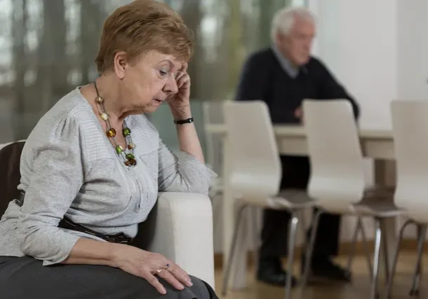 Worried elderly woman with husband in background.